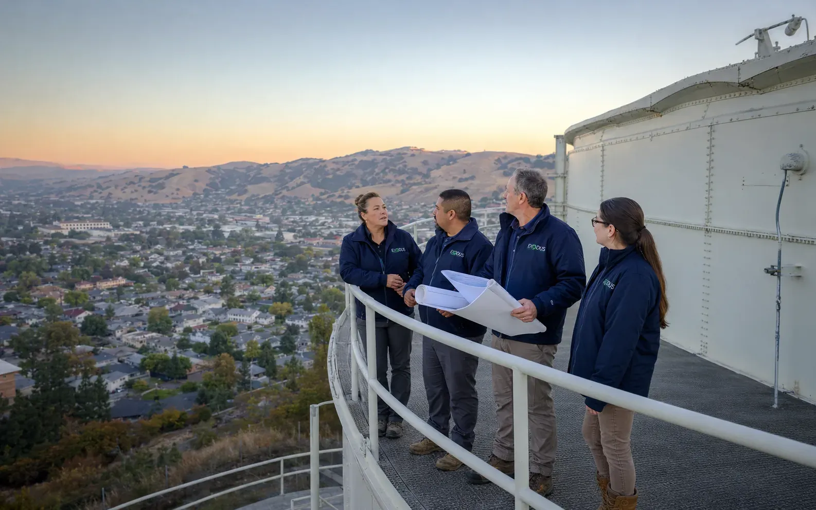 Four Exodus team members standing on a water storage tank catwalk above a town at sunset